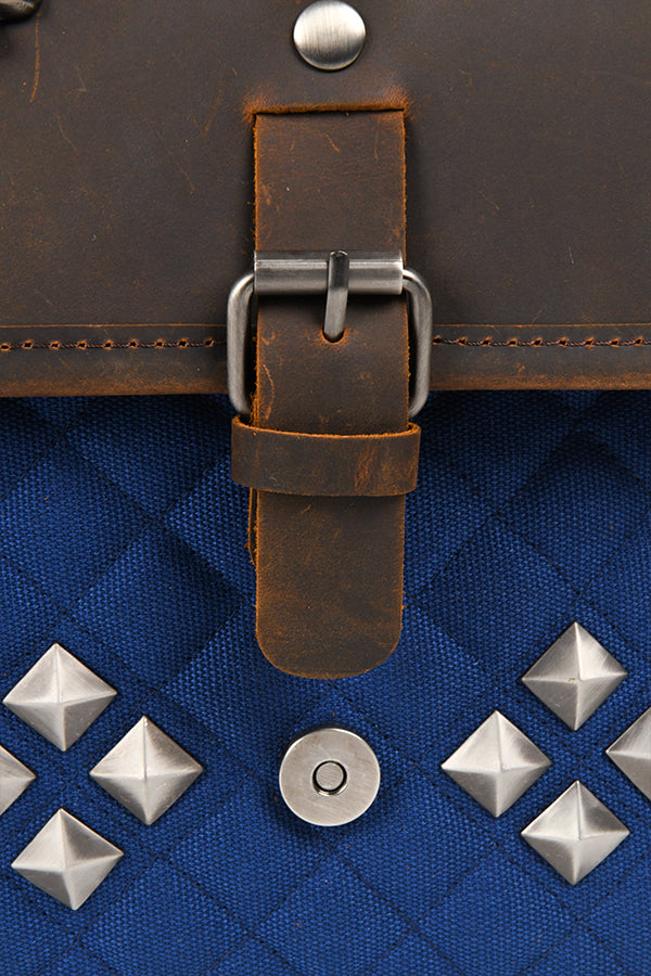 Detail shot of one of two front buckles on the leather flap, the button on the blue cotton canvas that it connects to, and the diamond-shaped studs flanking that button.