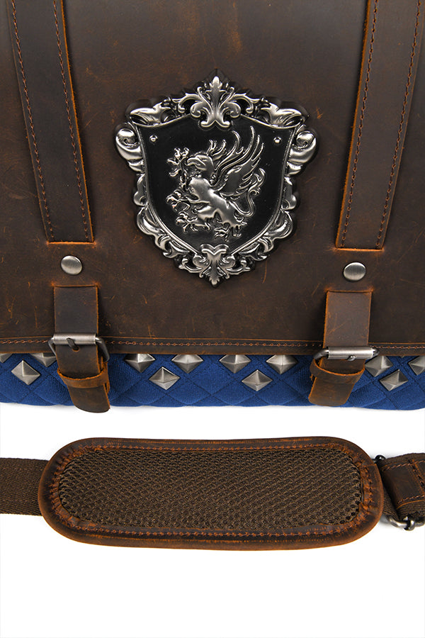 High angle detail shot of the front of the Dragon Age Grey Wardens Leather Bag, feat. the griffon crest, leather grain, matte finish metal accents, and the leather strap with a small, additional patch of leather stictched over it for support and shoulder comfort.