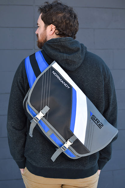 Image shows male model facing backwards wearing a gray hoodie under the Mass Effect SR2 Alliance Messenger Bag.  The product has colors and design elements from Commander Shepard's Normandy SR-2 that have quick release buckles on front flap and strap. It features top stitching details and brushed nickel hardware.