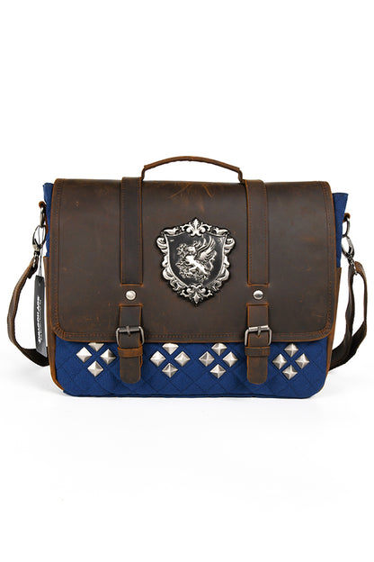 Image of the Dragon Age Grey Warden Leather Bag closed from the front, showing the contrast between the brown leather flap and the bag's blue cotton canvas exterior. The former features a pair of straps with buckles and a metal griffon crest. The latter features four groups of diamond-shaped studs arranged in a diamond pattern. These are in turn arranged in a row.