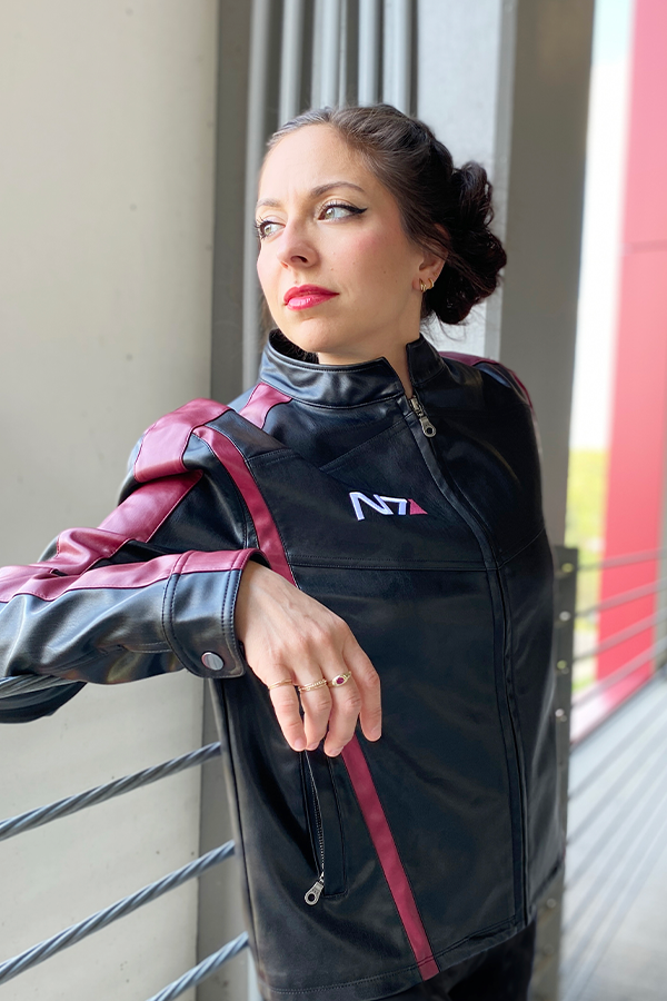 Image shows Mass Effect N7 Jacket Reimagined worn by female model while facing at an angle. According to The Art of Mass Effect, the red detail on the emblem symbolizes the human blood Shepard must sacrifice to stop Saren Arterius. The red stripe is also a historical reference to the red stripes commanders wore on early space missions to make them instantly recognizable.