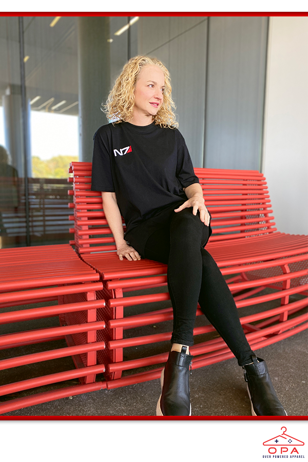 Image shows Mass Effect 3D Embroidered OPA T-Shirt worn by female model while sitting at a metallic bench with her legs crossed. N7 Day is a franchise appreciation day held every November 7 since 2012, where BioWare and fans offer support through content announcements, retrospectives, fan art, item sales, and more.