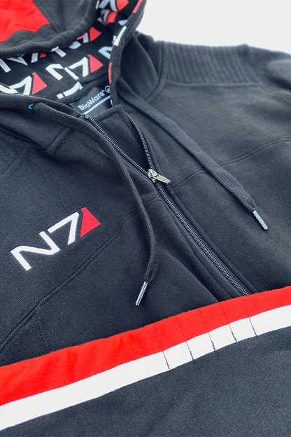 Image shows Mass Effect N7 Adult Onesie Reimagined laid flat with the upper part of the hoodie zoomed in. Product features a front zip closure and adjustable drawcord at rib waistband. Product also features right sleeve N7 stripes and a right chest N7 embroidered logo.