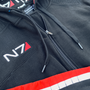 Image shows Mass Effect N7 Adult Onesie Reimagined laid flat with the upper part of the hoodie zoomed in. Product features a front zip closure and adjustable drawcord at rib waistband. Product also features right sleeve N7 stripes and a right chest N7 embroidered logo.