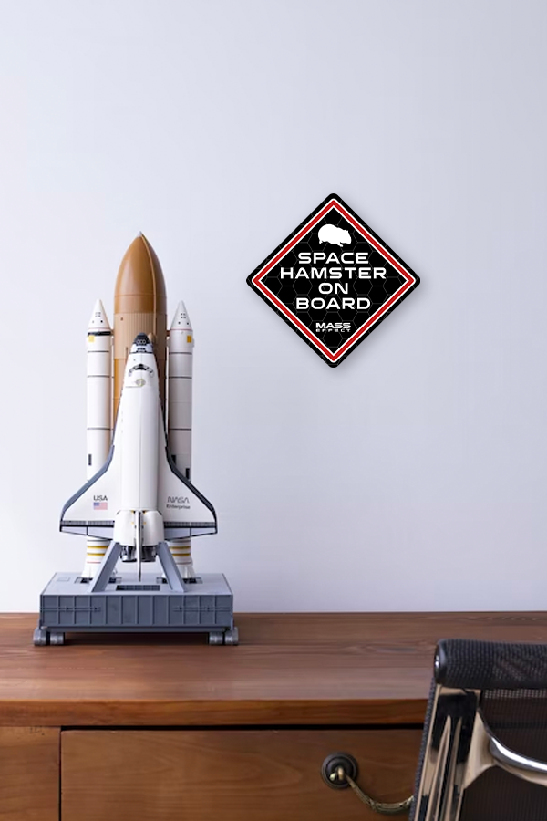Image shows Mass Effect Space Hamster on Board Vinyl Sticker on a wall beside a replica spaceship. You can add it to your laptop, wall, or door, and gift it to a fellow Mass Effect fan.