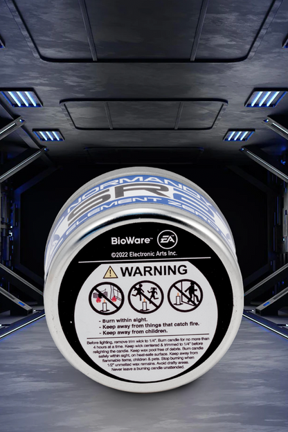Image shows Mass Effect Element Zero Scented Candle laid flat with its bottom facing front showing the Bioware and EA logos and warning instructions. Product features a 40 hour burn time and a scent of orange wood and cedar wood. 