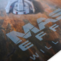 Image shows  Mass Effect Mysteries from the Future Lithograph upclose, laid flat at an angle, highlighting the Mass Effect text. Product features at least five surprises within the poster that will make this lithograph a great material for study and discussion.