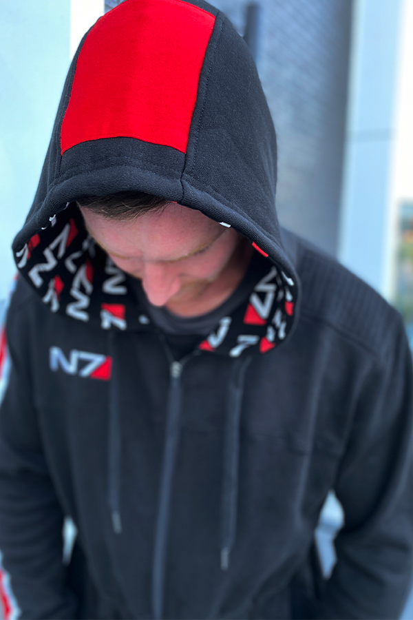 Image shows Mass Effect N7 Adult Onesie Reimagined worn by male model while looking down and the camera at a high point angle. Product features a color block hood details and adjustable drawcords at its hood.